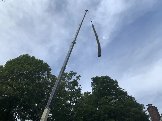Crane lifting a long, bent metal beam against a cloudy sky, with trees below.