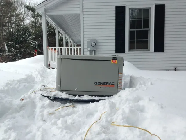 Gray generator sitting in snow beside a white house and porch with black shutters.