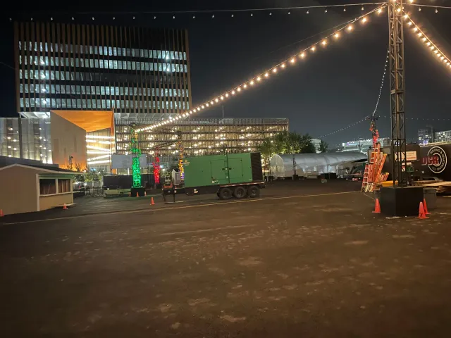 Night scene with string lights, buildings in the background, a green container on a trailer, and a small building to the left.