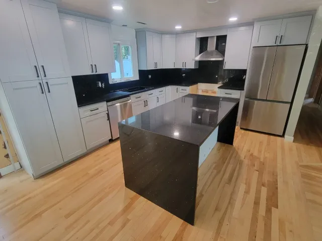 Modern kitchen with white cabinets, black countertops, stainless steel appliances, and a central black island, with light wood flooring and a window above the sink.