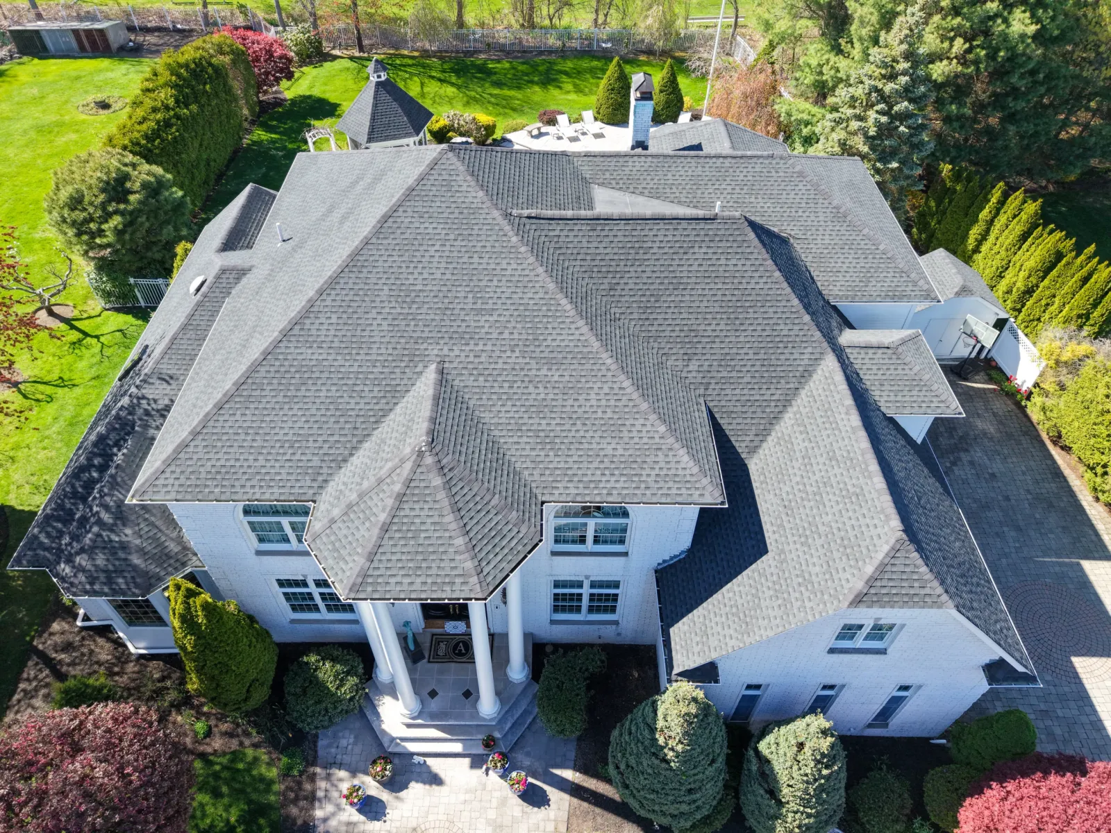 Aerial view of a large, elegant house with a gray, multi-gabled roof, white exterior walls, a grand entrance with columns, and a well-manicured garden and driveway surrounding the property.