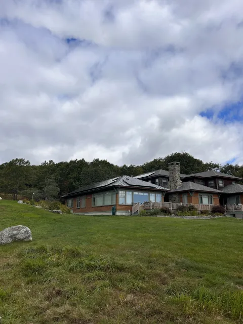 A large house with a stone chimney, wooden exterior, and a large front window, situated on a grassy hill with trees in the background under a partly cloudy sky.