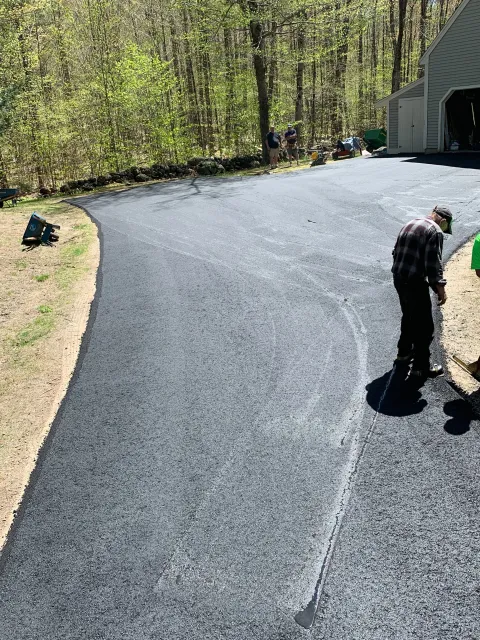 People working on freshly paved asphalt driveway in a wooded area, with tools and equipment nearby.