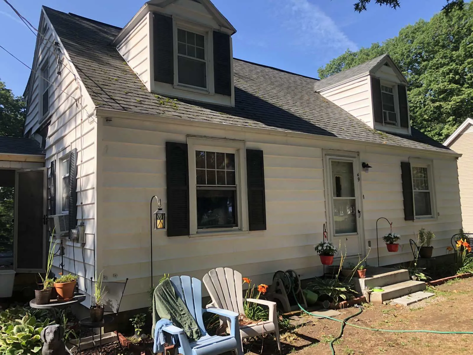 A white, one-and-a-half-story house with two dormers, black shutters, and blue and white chairs on the lawn.