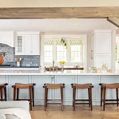 Bright white kitchen with blue island, bar stools, and a windowed dining nook under exposed wood beams