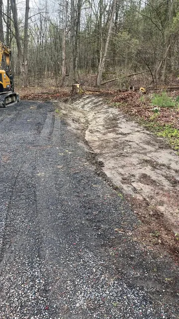 Gravel road beside muddy, leaf-covered edge in a wooded area, with part of a yellow machine at left.
