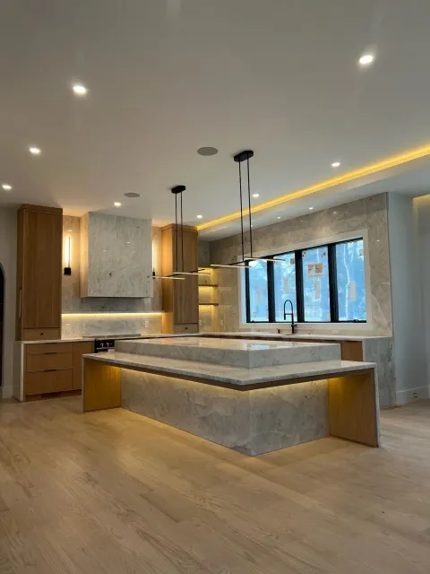 Modern kitchen with marble countertops, wooden cabinetry, an island, black pendant lights, large window, and warm lighting.