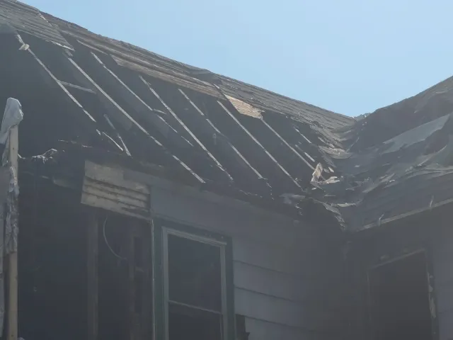 Damaged roof of a house with missing shingles and burnt wood, exterior walls with visible siding, two windows, and a clear blue sky.