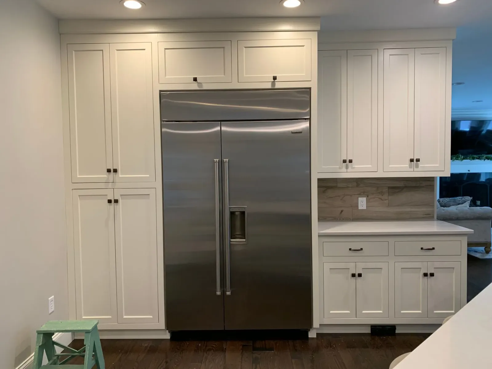 A stainless steel refrigerator sits between white cabinet units in a kitchen with hardwood floors and a tiled backsplash.