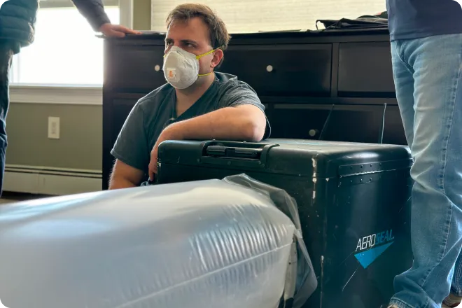 A person wearing an N95 mask sits on the floor next to a large, black AeroSeal machine with plastic tubing attached; another person stands nearby.