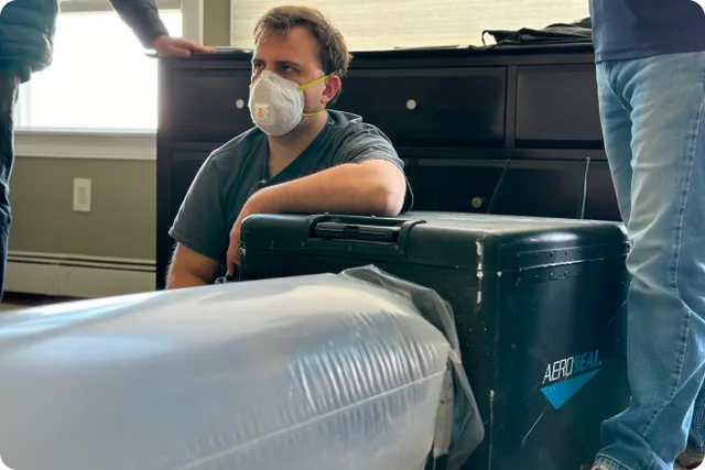 A person wearing an N95 mask sits on the floor next to a large, black AeroSeal machine with plastic tubing attached; another person stands nearby.