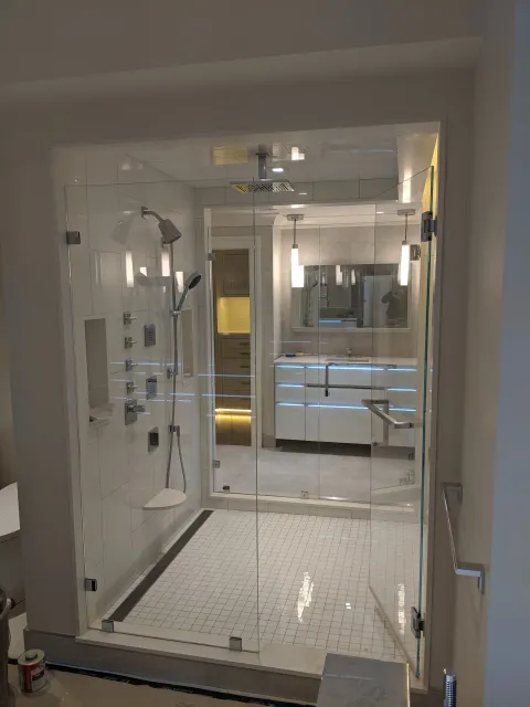A modern walk-in shower with glass walls, multiple shower heads, and a built-in bench, with a view into a well-lit bathroom and vanity area.