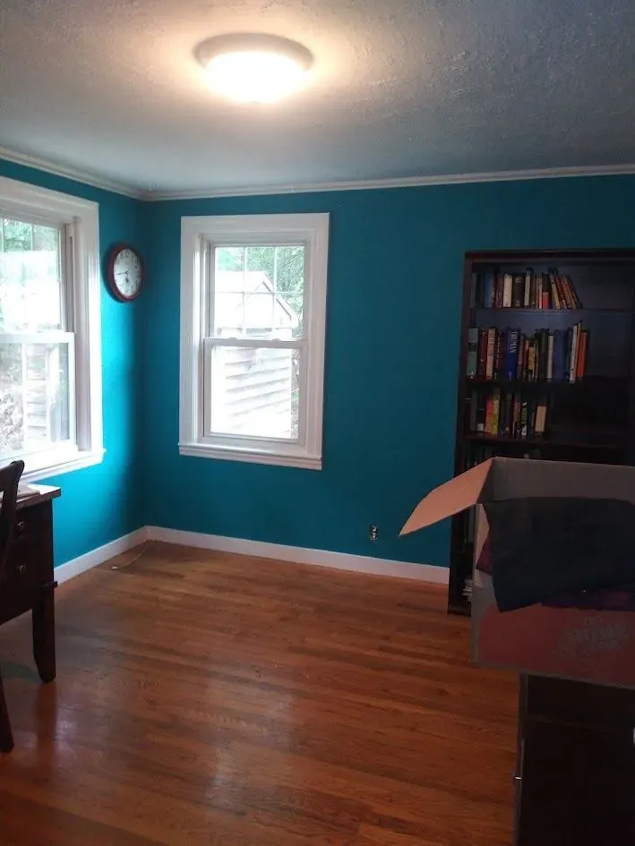 Empty room with teal walls, hardwood floors, two windows, ceiling light, and a bookshelf with a covered chair.