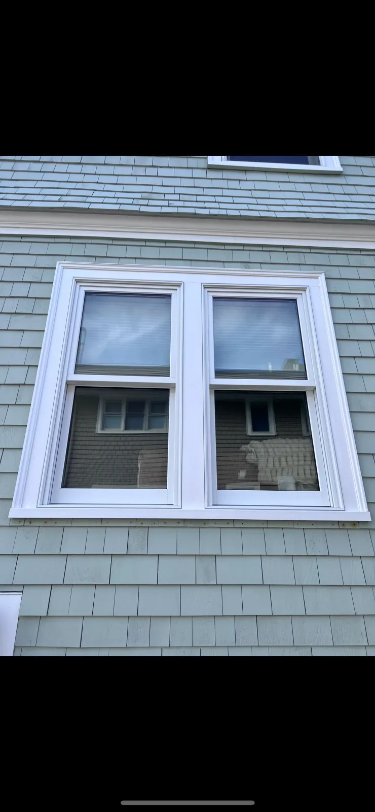 Close-up of a double-hung window with white trim on a light green shingled house, with reflections of the sky and nearby house in the glass panes.