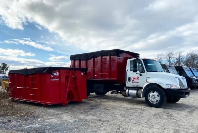 Red dump truck with a red dumpster trailer parked on a gravel lot under a cloudy sky