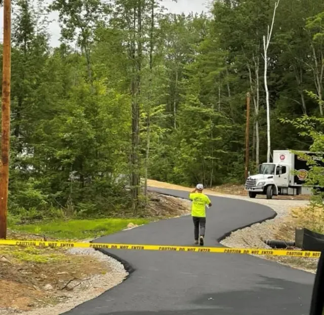 A person in a bright yellow shirt walks down a freshly paved, winding asphalt path bordered by trees and caution tape.