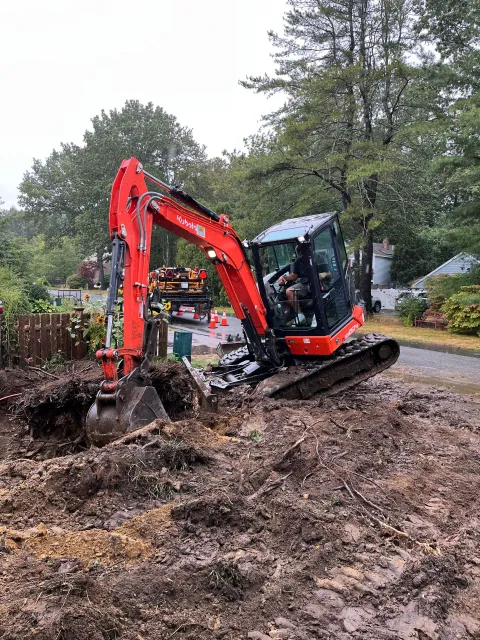 Red excavator digging in muddy soil near a road and fence, with a person inside.