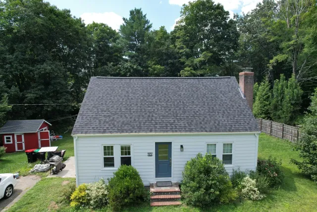 White house with dark roof, blue front door, and green yard beside a red shed, surrounded by trees.