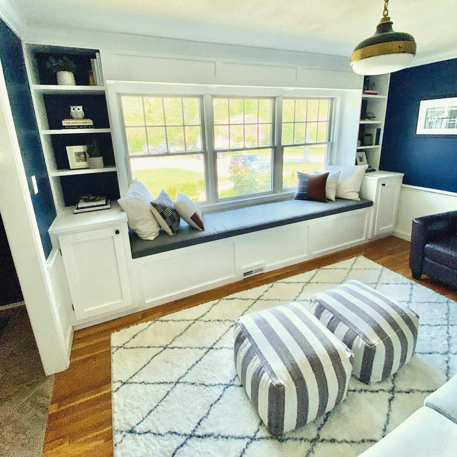 Living room with a large window seat under a window, built-in shelves with books and decor, a black accent wall with a framed picture, a white rug with black diamond pattern, and two striped ottomans.