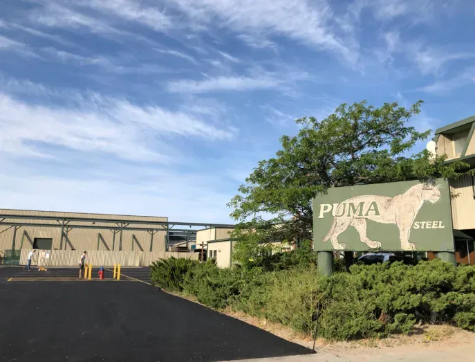 Industrial site with a Puma Steel sign beside a paved road and low buildings under a blue sky