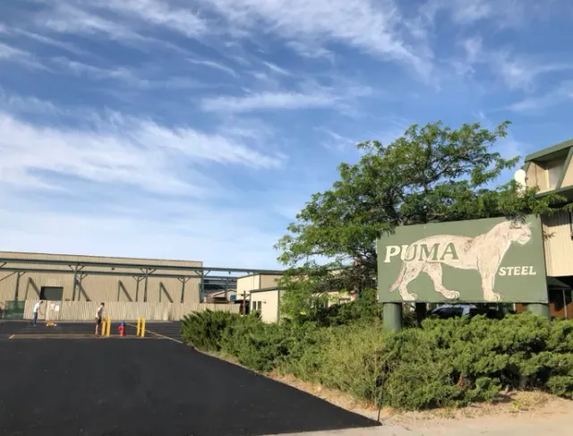 Industrial site with a Puma Steel sign beside a paved road and low buildings under a blue sky