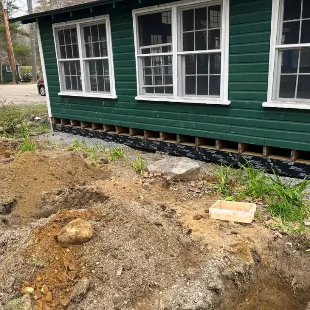 Green house with white-trimmed windows beside a dirt excavation area and exposed foundation vents