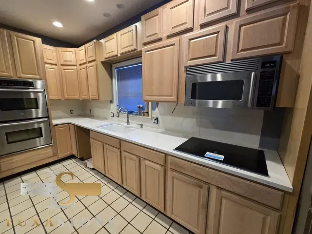 A kitchen with light wood cabinets, white countertops, a stainless steel microwave, and a double oven.