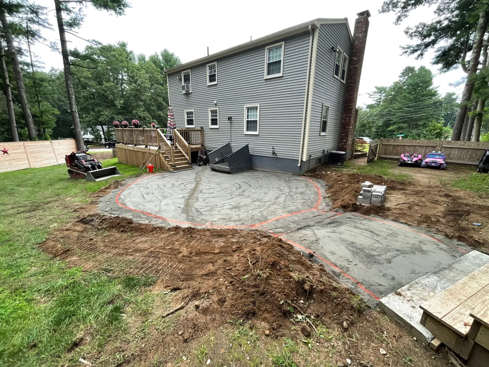 Backyard with construction in progress, showing a large circular groundwork area, a wooden house deck, small machinery, and piles of materials.
