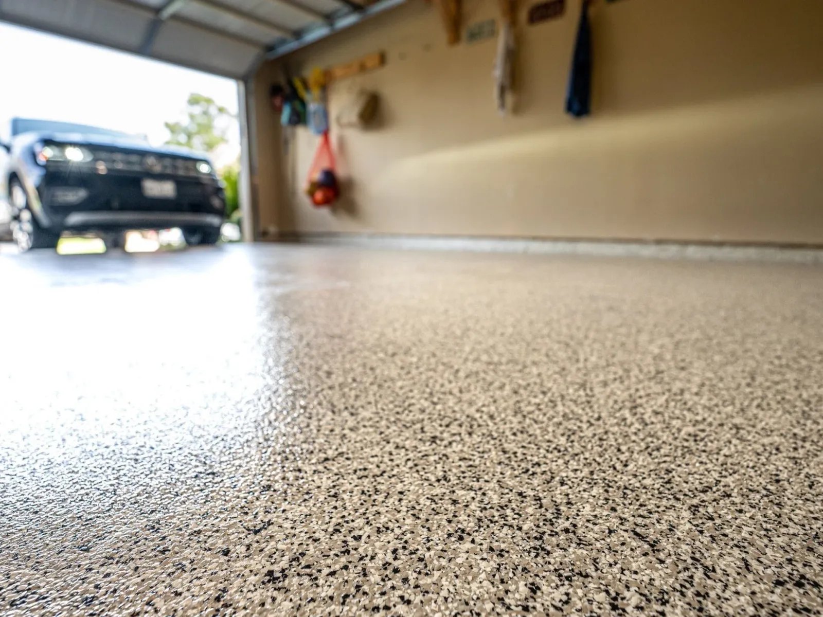 Inside view of a garage showing a textured floor, with a black vehicle parked outside and tools hanging on the wall.