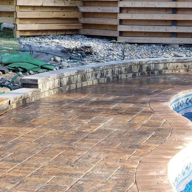 Stamped concrete patio with a stone retaining wall alongside a backyard swimming pool and wooden privacy fence.