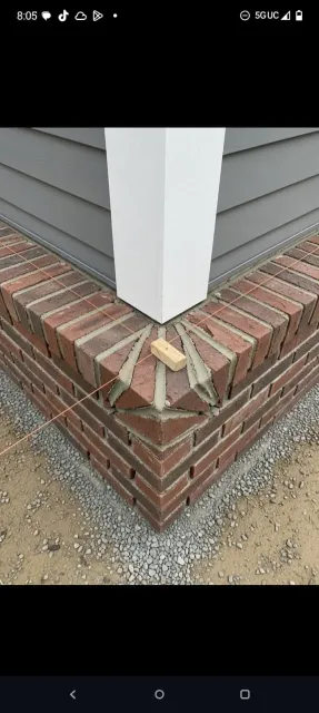 Corner of a house with brick foundation and white trim, showing a damaged piece at the base.