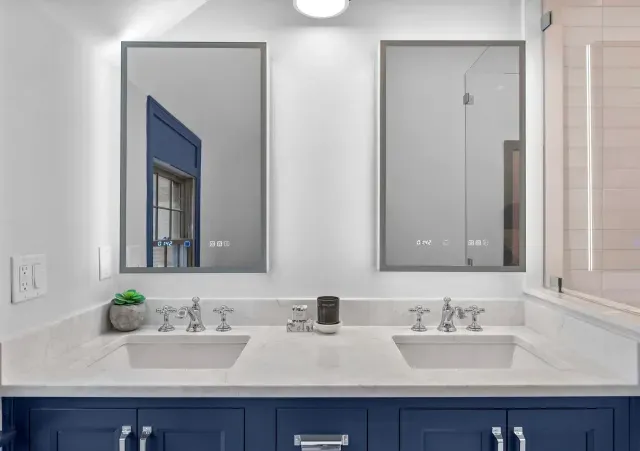 Bright bathroom vanity with double sinks, two mirrors, and blue cabinets under a window.