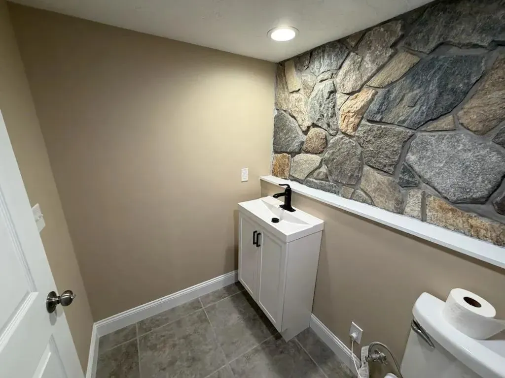Small bathroom with white vanity, stone accent wall, and toilet paper on a side shelf.