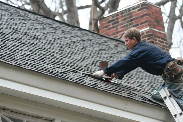 Roofer on a ladder repairing shingles next to a brick chimney and white trim.