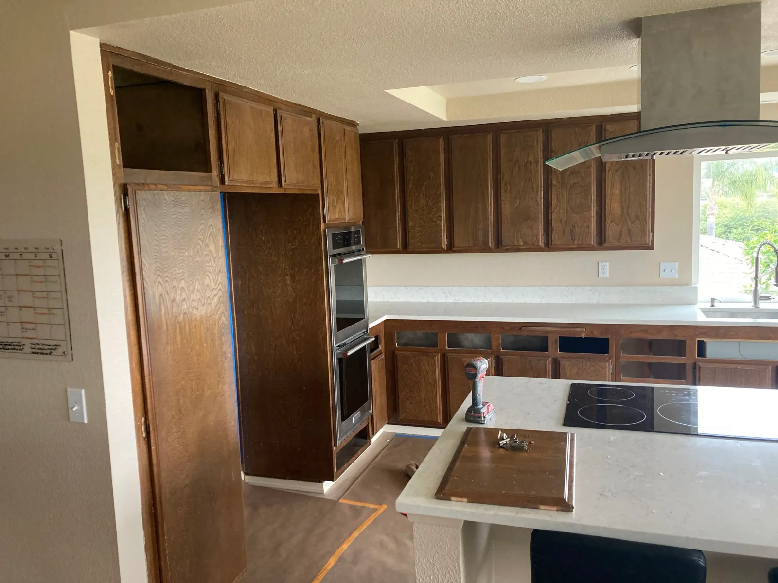 A kitchen with wooden cabinets, a built-in oven, a stove on the island, and a window with greenery outside.
