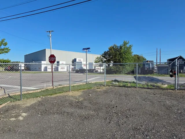 Chain-link fence beside a gravel lot, with a warehouse building and stop sign under a clear blue sky.