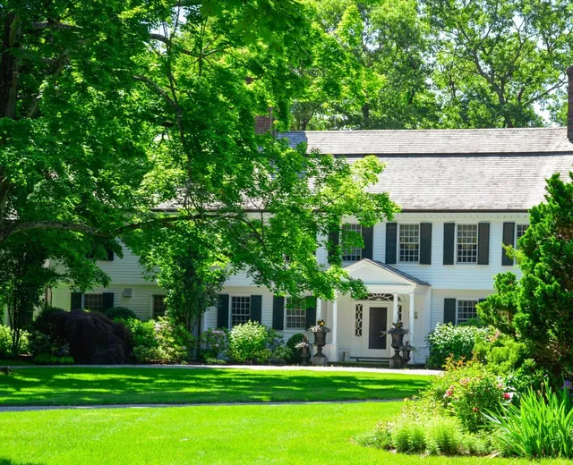 This is a two-story, white colonial house with green shutters surrounded by lush greenery, manicured lawn, and well-maintained garden beds on a sunny day.