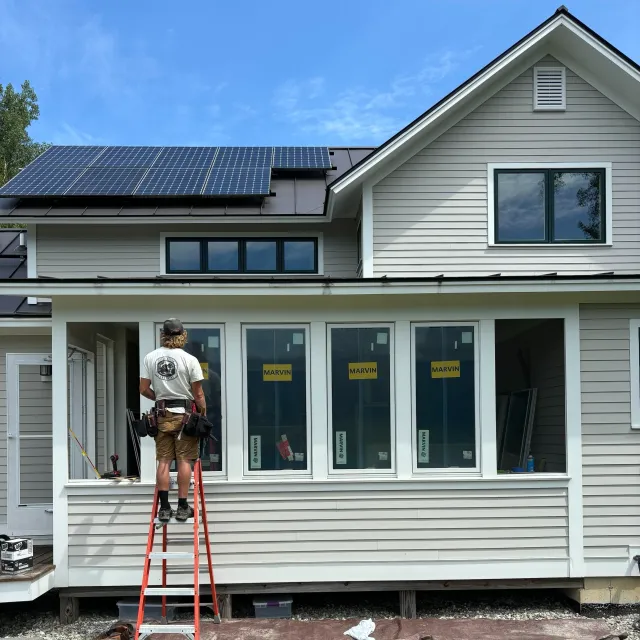 Construction worker installing windows on a house porch with existing siding and solar panels on the roof under a blue sky.