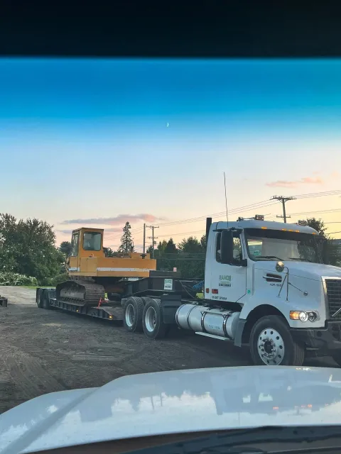 A white semi-truck hauling a yellow excavator on a flatbed trailer on a dirt lot at dusk.