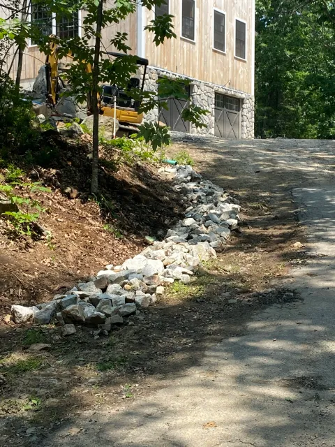 A construction site beside a partly built wooden house with garage doors, showing a stone-lined drainage ditch along the dirt road.