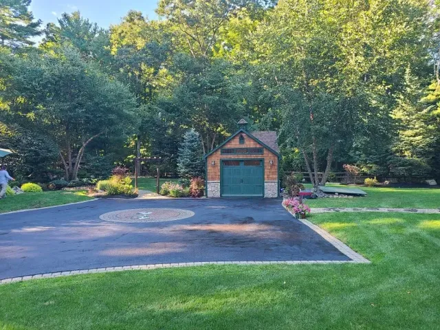 A small, shingled shed with green double doors sits at the end of a paved driveway in a wooded backyard.