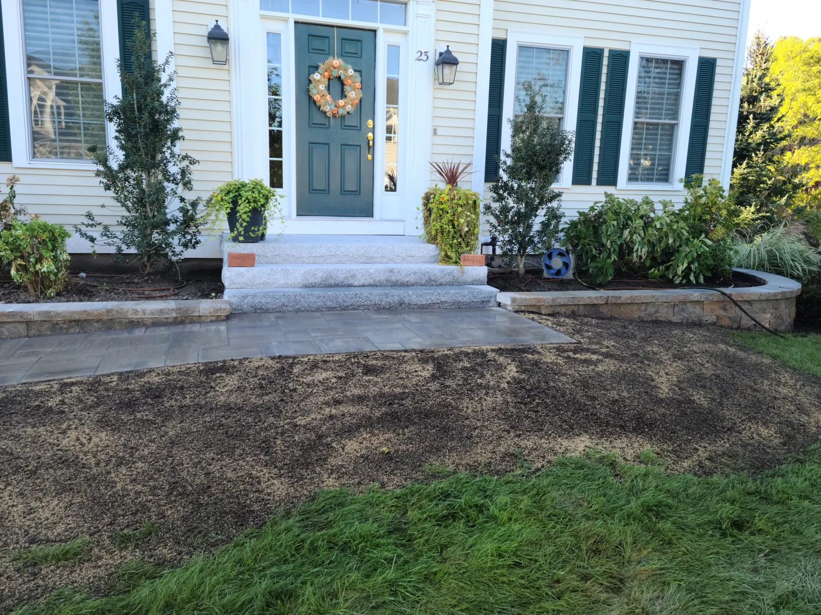 Front porch of a house with a green door, decorated with a wreath. There are steps leading to the door, with landscaped flower beds on each side, plants, and bushes. A stone and paved pathway is in front of the stairs, with some freshly laid soil and grass extending outward.