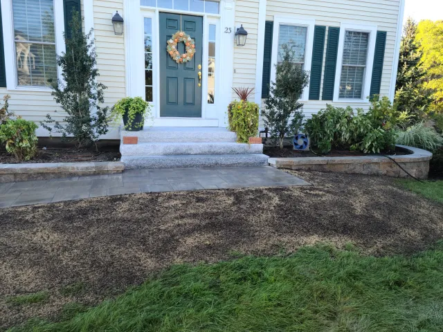 Front porch of a house with a green door, decorated with a wreath. There are steps leading to the door, with landscaped flower beds on each side, plants, and bushes. A stone and paved pathway is in front of the stairs, with some freshly laid soil and grass extending outward.