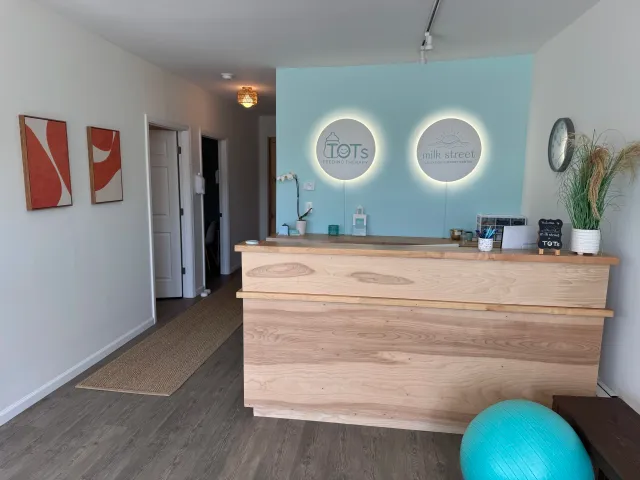 The reception area of a breastfeeding support and therapy center called TOTS feeding therapy and Milk Street Lactation Support Center, with a wooden reception desk, wall decorations, and a large clock, plus a blue exercise ball in the corner.