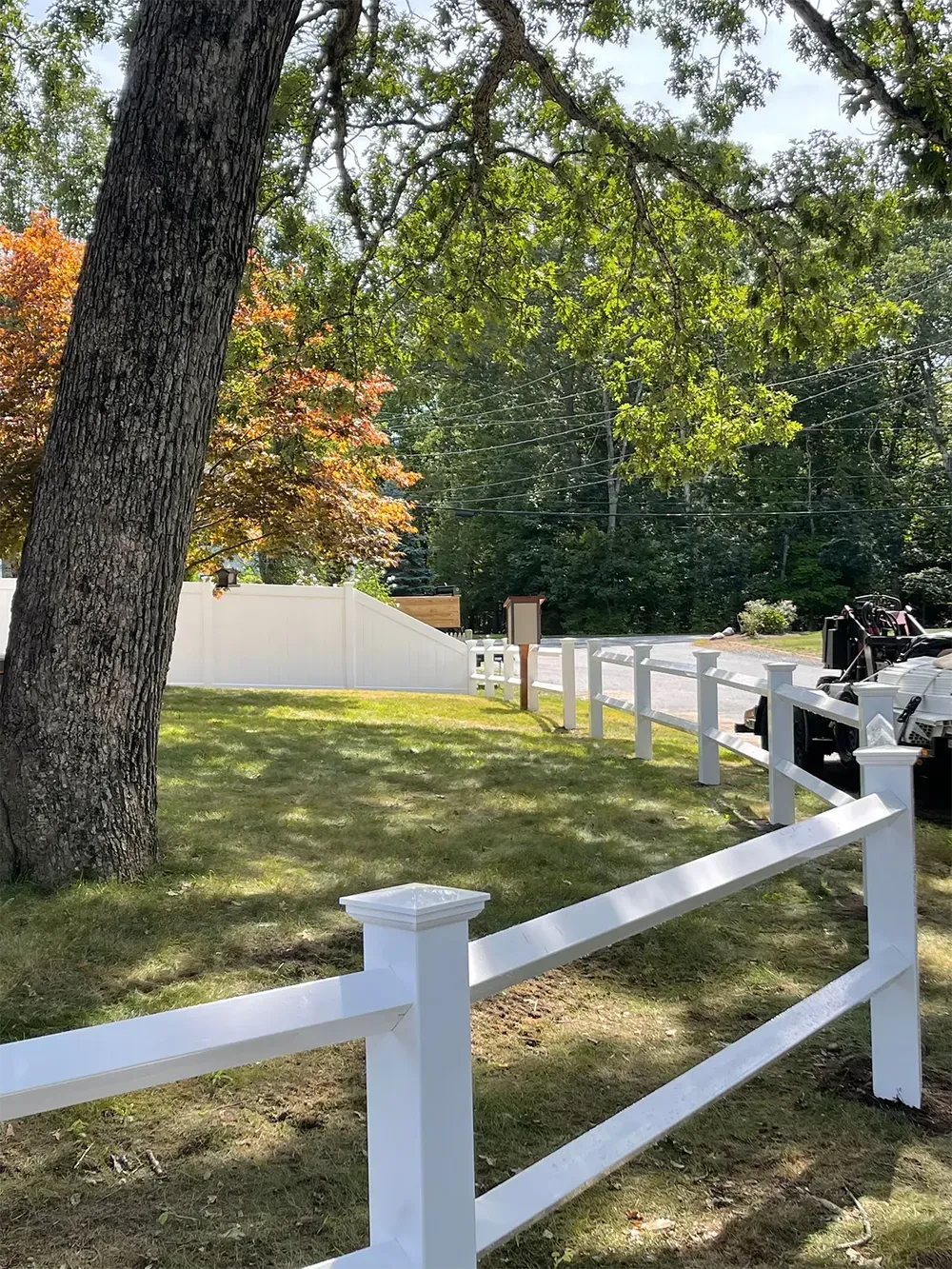 White fence bordering a grassy area, leading to a driveway with a gate. Trees with autumn foliage.