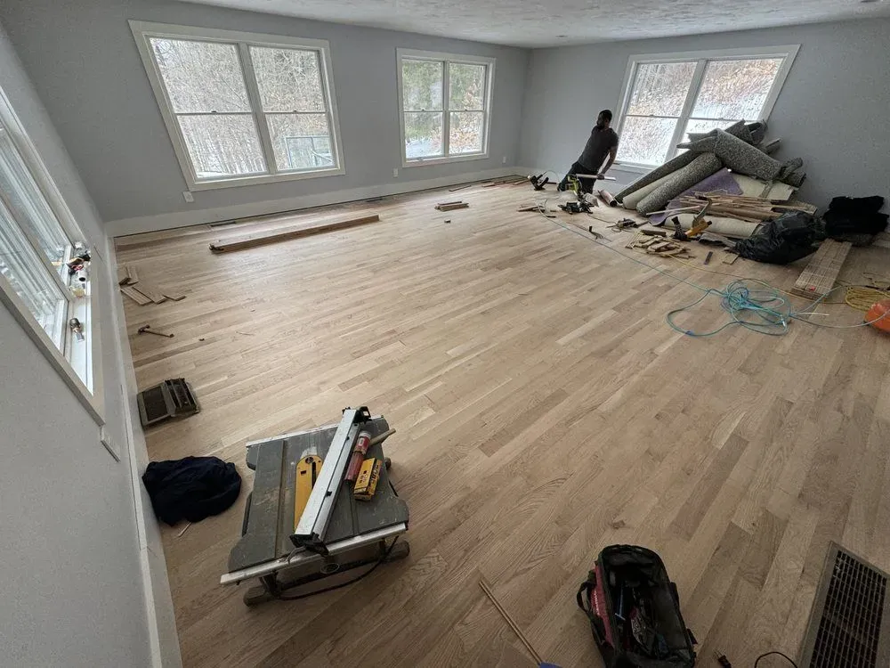 Room with light wood floor, windows, and construction tools. A person sits near a pile of materials.