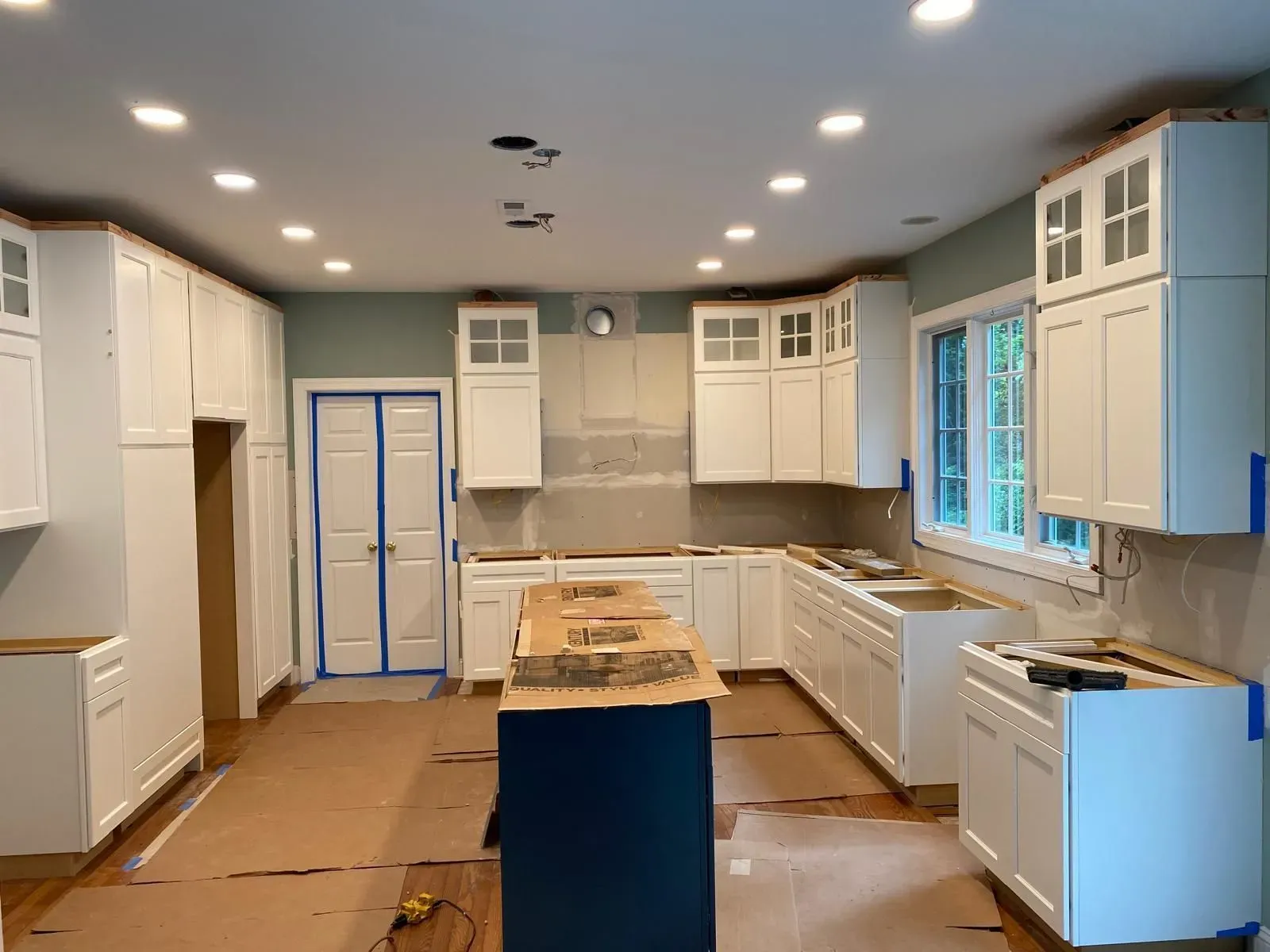 Renovated kitchen with white cabinets, blue island, and unfinished flooring under bright recessed lights