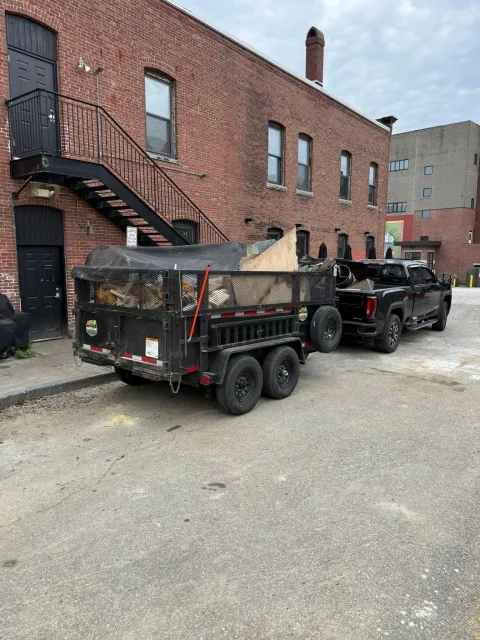 Black dump trailer hitched to a pickup in a brick alley beside a building with an exterior staircase