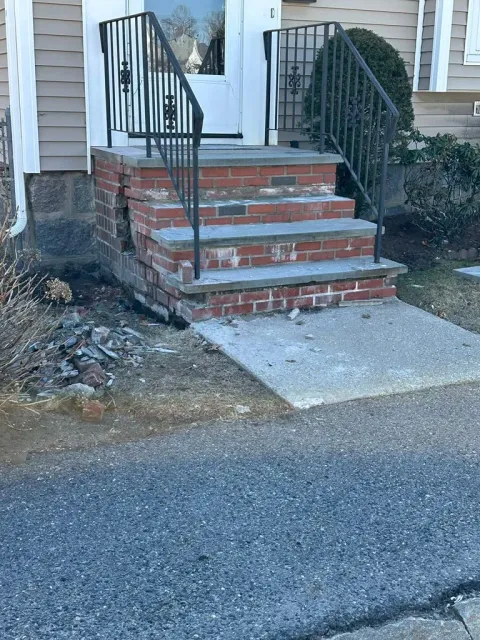 Front steps to a house with brick stairs, black railings, and a concrete landing beside a driveway.