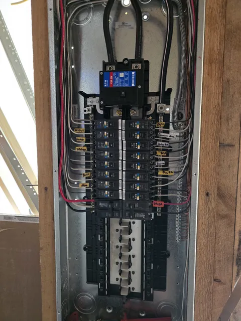 Electrical panel with multiple circuit breakers and red and black wires, housed in a metal enclosure. The panel is mounted on a wooden structure.